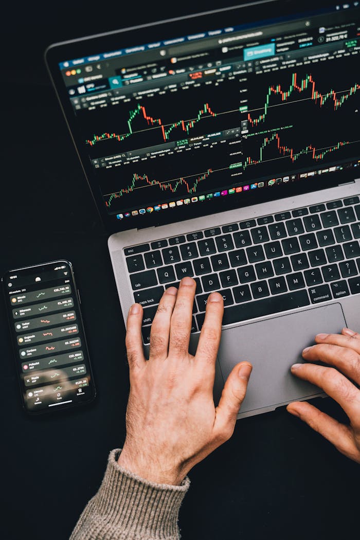 Hands typing on a laptop with cryptocurrency charts and smartphone display in view, focusing on digital finance.